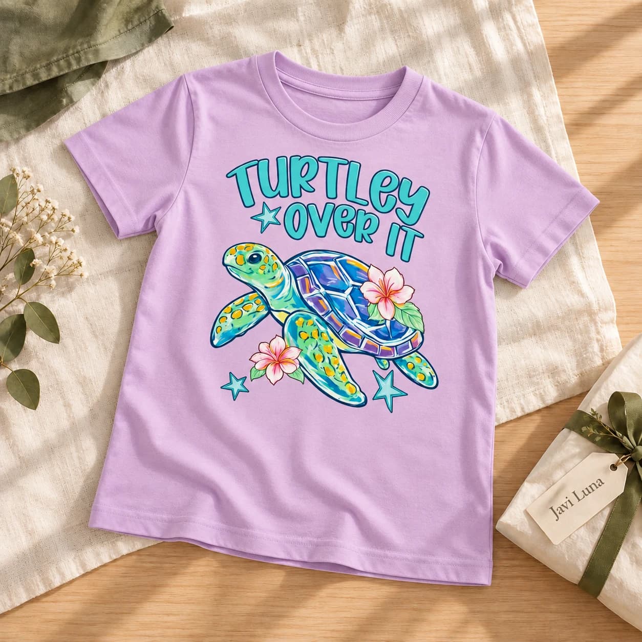The lavender Turtley Over It tee laid on linen with eucalyptus and a Javi Luna gift box